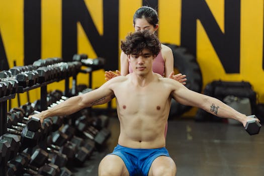 A shirtless man lifting dumbbells with a female trainer's guidance in a gym setting.
