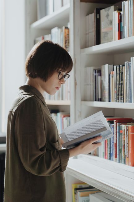 A young woman wearing glasses reads a book in a well-lit library.