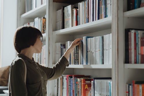 Man in Brown Long Sleeve Shirt Holding Book in White Wooden Book Shelf