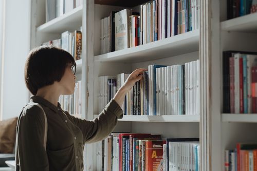 Free A woman browsing a bookshelf in a modern library setting, exploring various literature options. Stock Photo