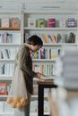 一位女士在书店里翻看书籍，手里拎着一个装着橙子的环保袋