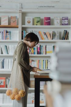 一位女士在书店里翻看书籍，手里拎着一个装着橙子的环保袋