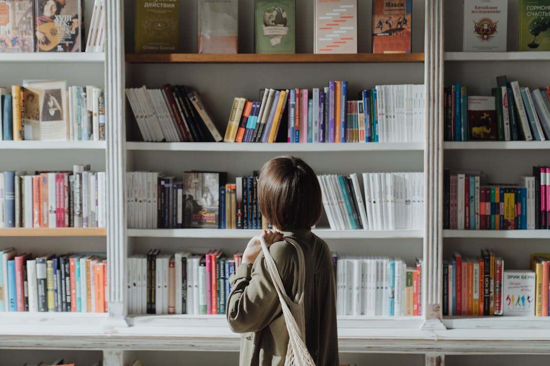 Free Woman in Beige Coat Standing Near White Wooden Book Shelf Stock Photo