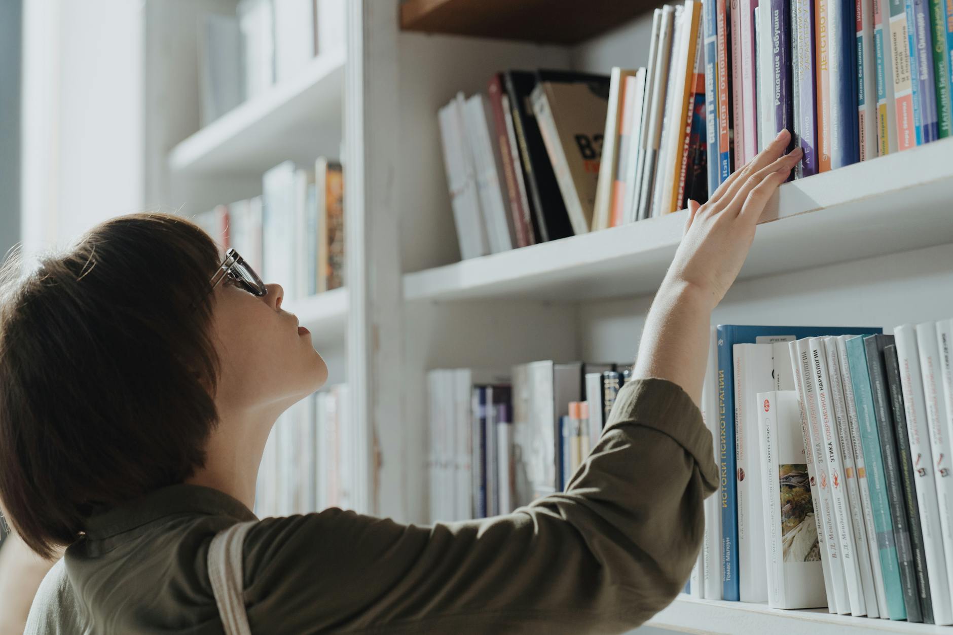 A woman wearing glasses browsing books on a library shelf, seeking knowledge and information.