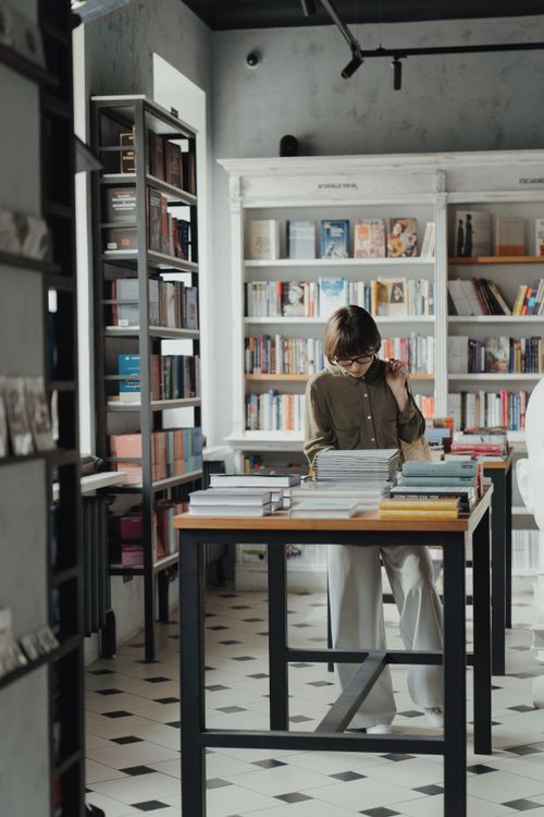 Free A young woman in glasses reads in a modern bookstore with stacks of books around her. Stock Photo