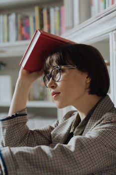 图书馆里，一位戴眼镜的女人头上顶着一本书，正全神贯注地思考