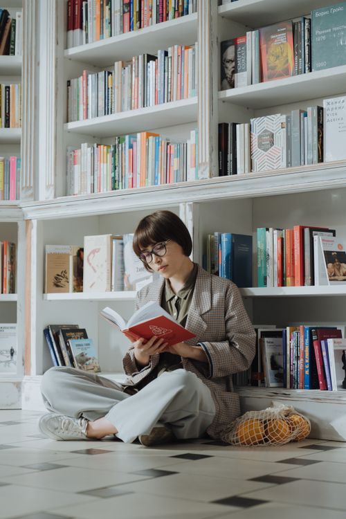Free A young woman sitting on the floor of a library, engrossed in a book, surrounded by shelves filled with colorful books. Stock Photo