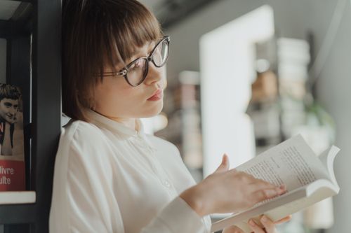 Free Young woman in glasses and white shirt reading a book in a bookstore setting. Stock Photo