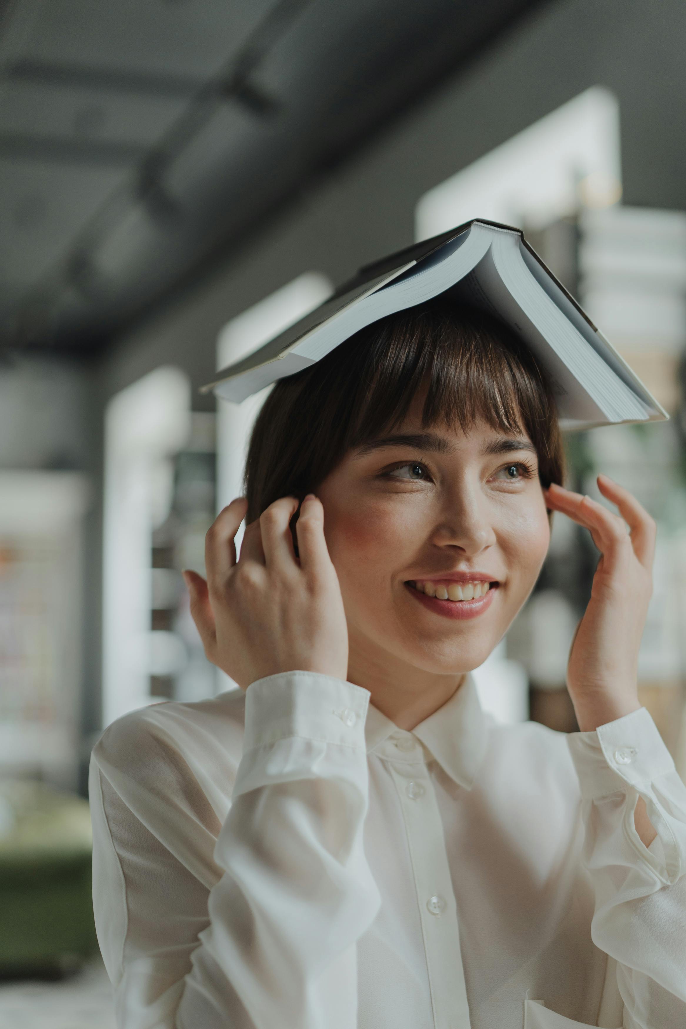 一位微笑的女人在图书馆里，把一本打开的书顶在头上，享受着休闲阅读