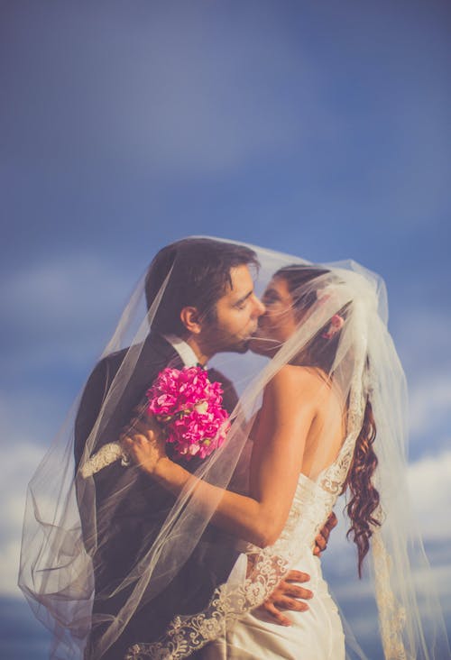Back view newlywed couple in chic wedding gowns kissing under veil while standing in nature on clear summer day
