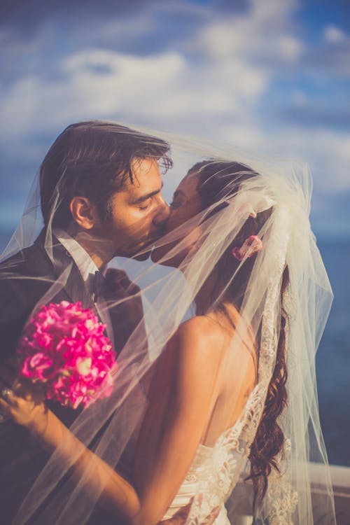 Free Side view loving newlywed couple wearing luxurious wedding gowns standing in nature under translucent veil while embracing and kissing gently with eyes closed Stock Photo