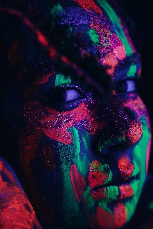 Crop female with shimmer and colorful neon paints on face glowing under ultraviolet light