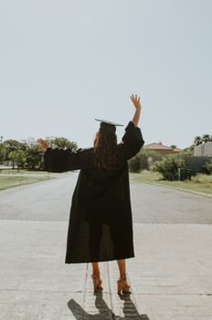 一位穿着毕业礼服的妇女在户外举起双手庆祝成功