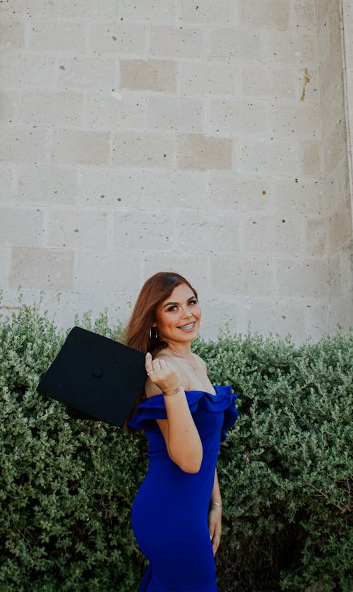 Cheerful young graduate in elegant vibrant blue dress looking at camera with bright smile while showing black square academic cap next to hedge and brick wall of building
