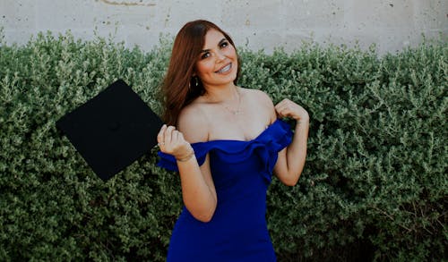 Cheerful female graduate holding academic cap next to brick wall