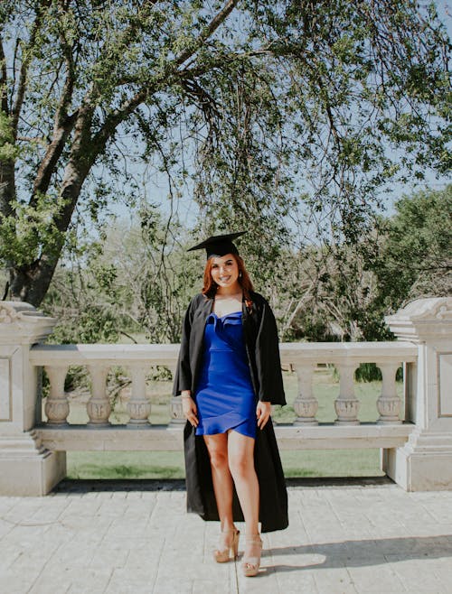 Full body of smiling young graduate in short blue dress and black square academic cap looking at camera with bright smile