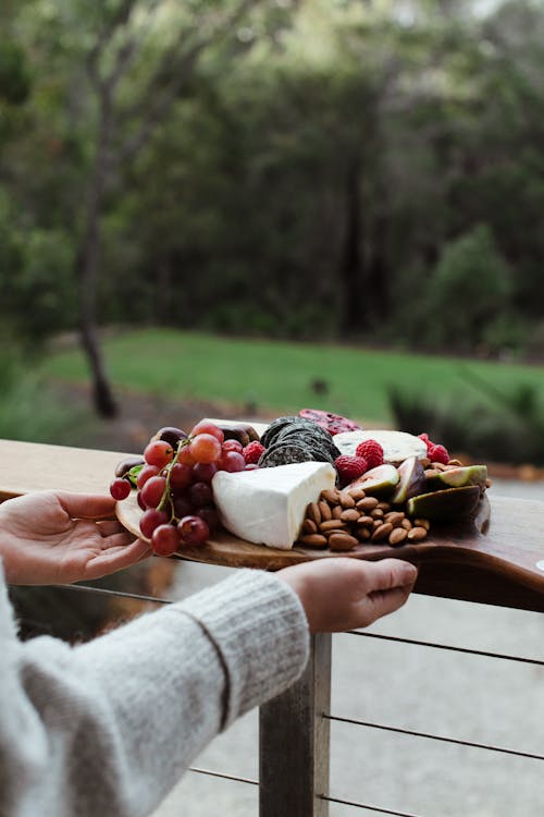 Crop anonymous female in sweater holding delicious cheese platter served with ripe juicy grapes and almonds while standing on veranda against blurred summer forest