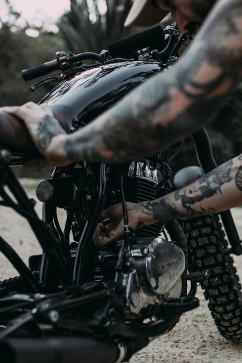 Crop unrecognizable tattooed male biker checking engine on black metal motorbike before riding