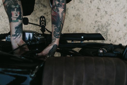 Top view of crop unrecognizable tattooed male biker repairing engine of motorbike before riding