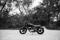 一辆黑色和白色的金属摩托车停放在靠近沙地道路的森林里，周围是落叶树木和茂盛的灌木丛