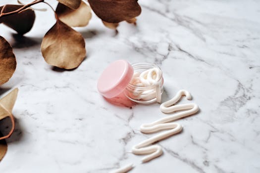 Close-up of skincare cream in a pink glass jar on a marble surface surrounded by leaves, ideal for beauty and wellness themes.