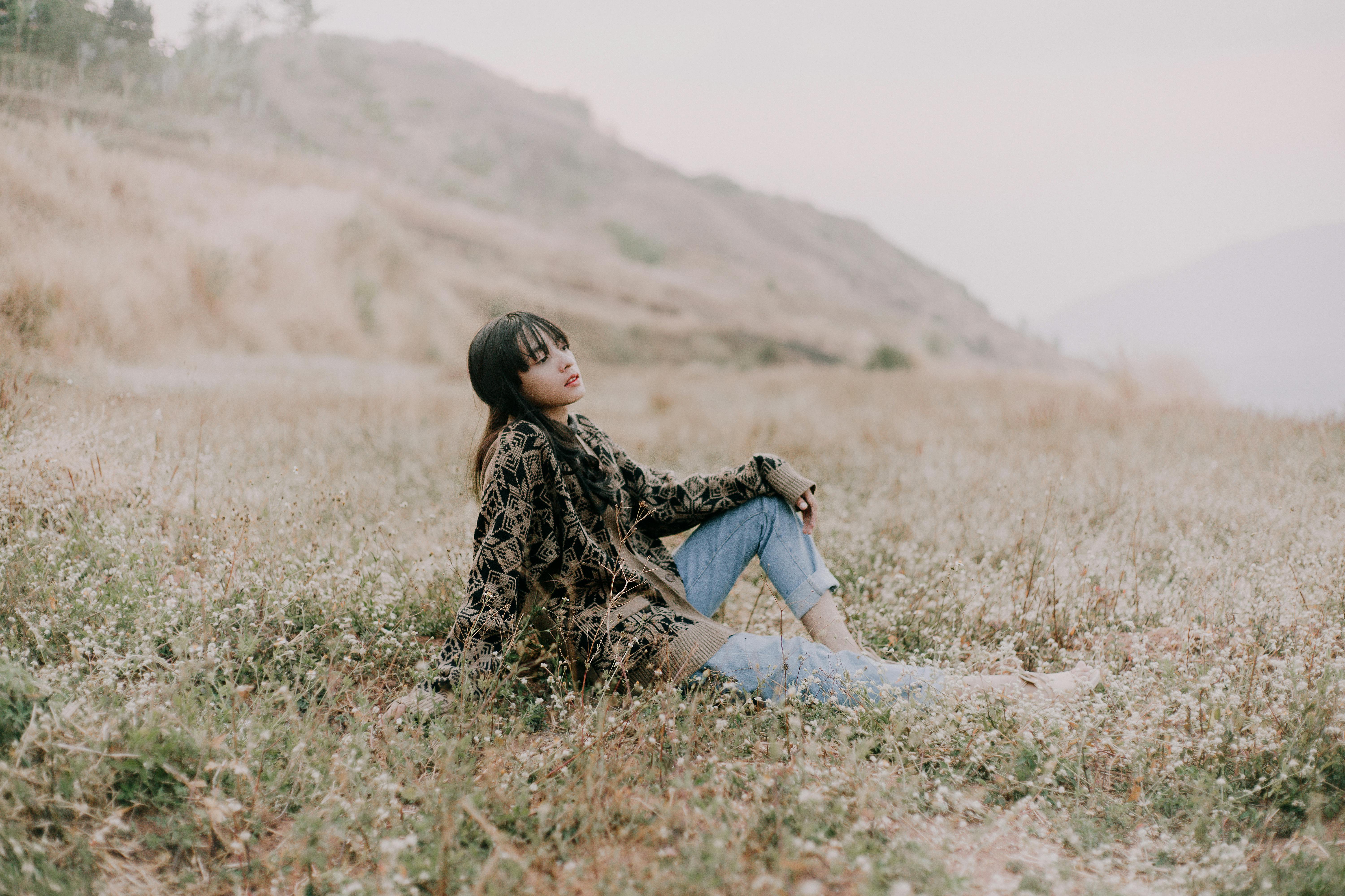 An Asian woman sits peacefully in a grassy field wearing a cozy sweater.