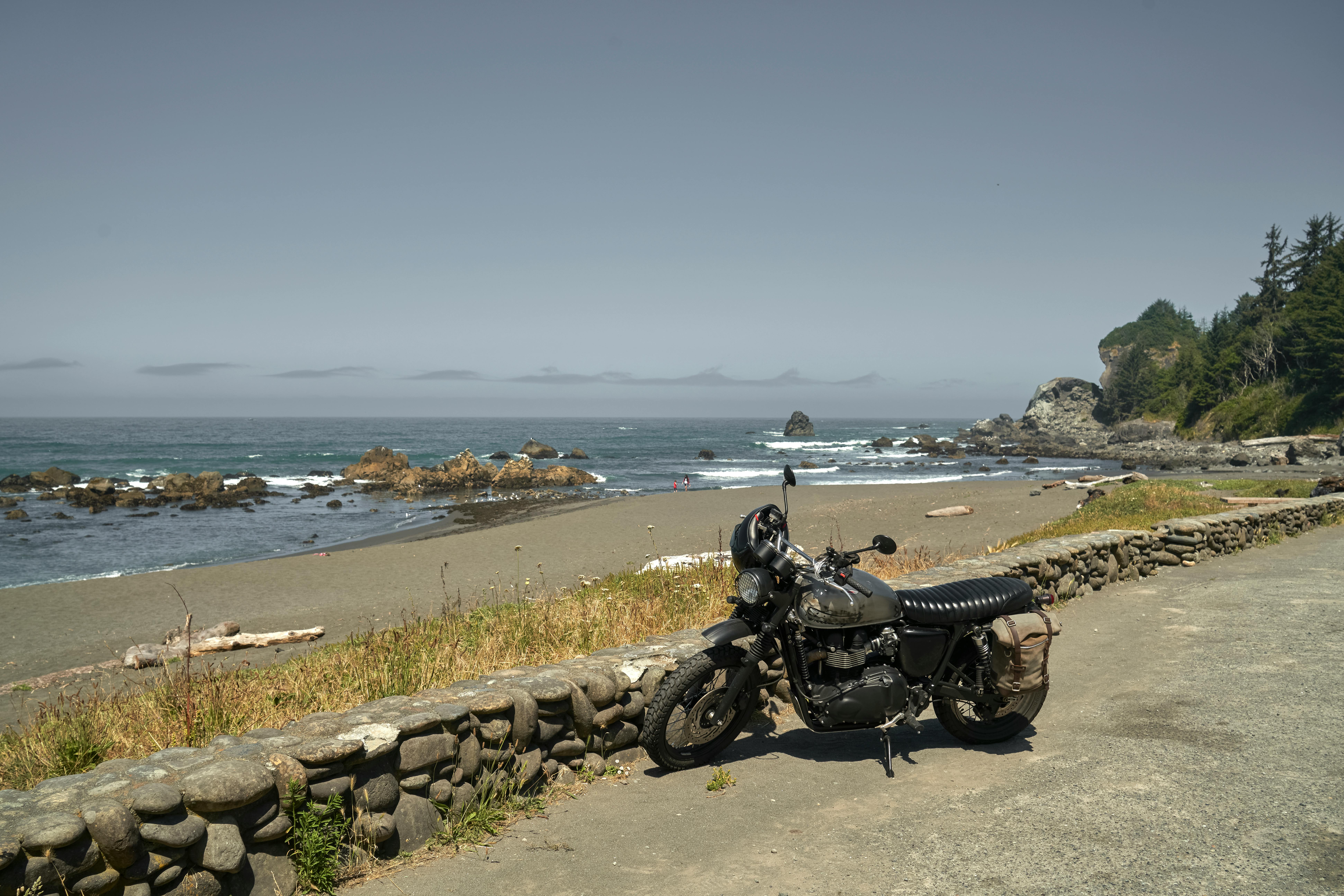 A classic motorcycle parked along a rocky beach with ocean waves and clear summer skies.