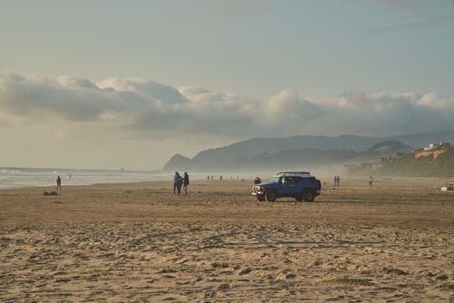 Free Blue SUV and beachgoers on a sandy beach with distant mountains under cloudy sky. Stock Photo