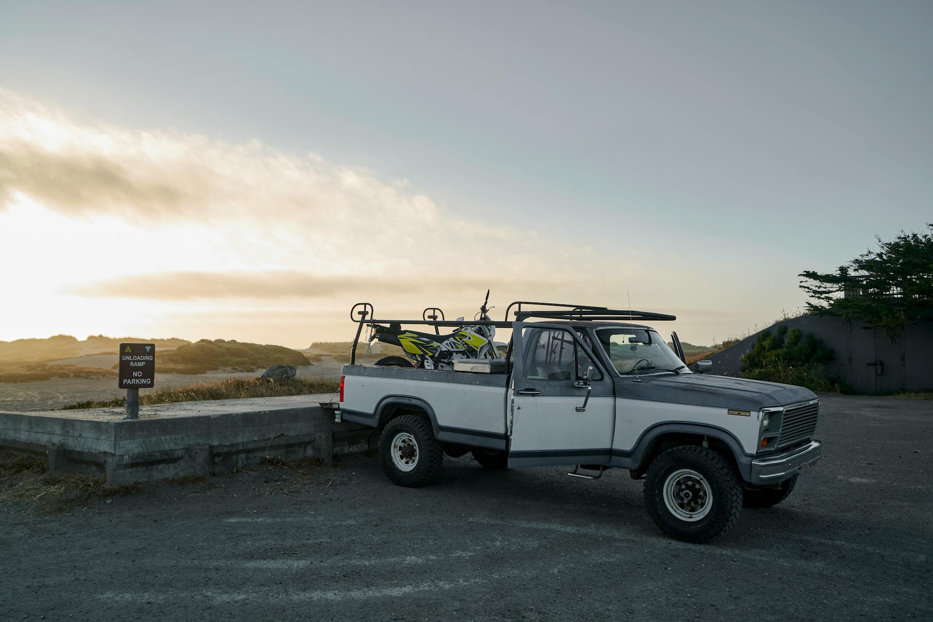 A classic pickup truck parked at a scenic spot during twilight with a dirt bike loaded on the back.