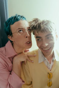 Two teenagers with colorful hair and eyewear make funny faces in a playful photobooth setting.