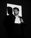 Black and white of brutal male in elegant suit standing in dark studio under light while casting shadow on wall and looking away