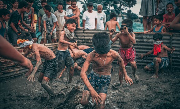 Kids play a traditional mud game, blindfolded and joyful, surrounded by onlookers.