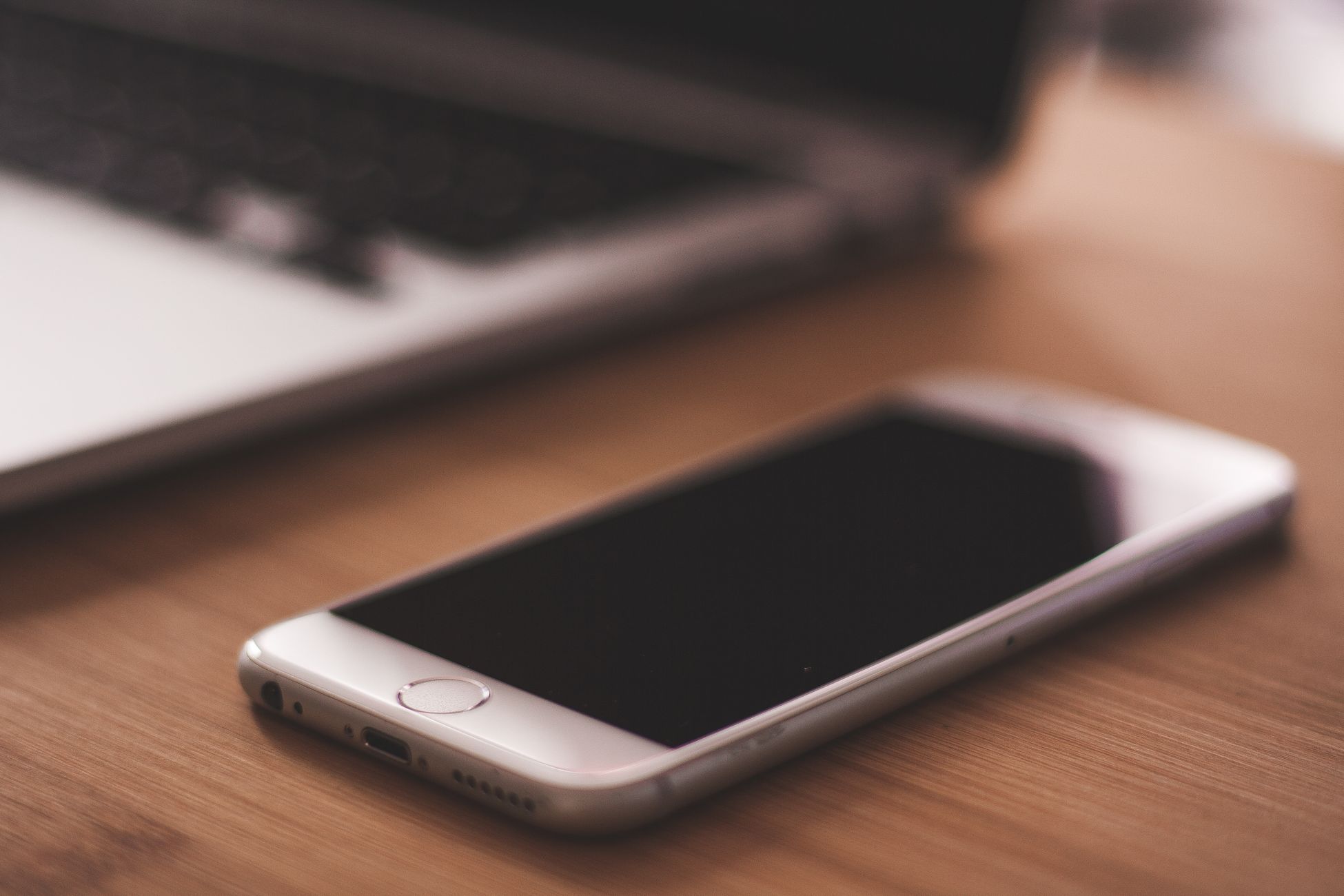 A smartphone lies on a wooden table next to a blurred laptop in a cozy indoor setting.