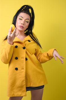 一位穿着黄色外套的年轻女子，手持柠檬，在黄色背景前自信地摆出姿势