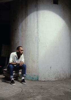 一名棕色短发、穿着白衬衫和牛仔裤的严肃少数族裔男性，坐在古老的白色混凝土墙旁边