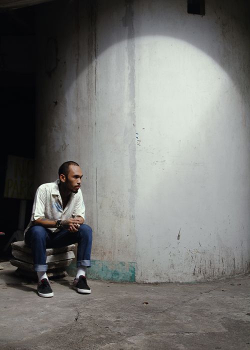 Free Serious ethnic male with short brown hair in white shirt and jeans sitting near old white concrete wall Stock Photo