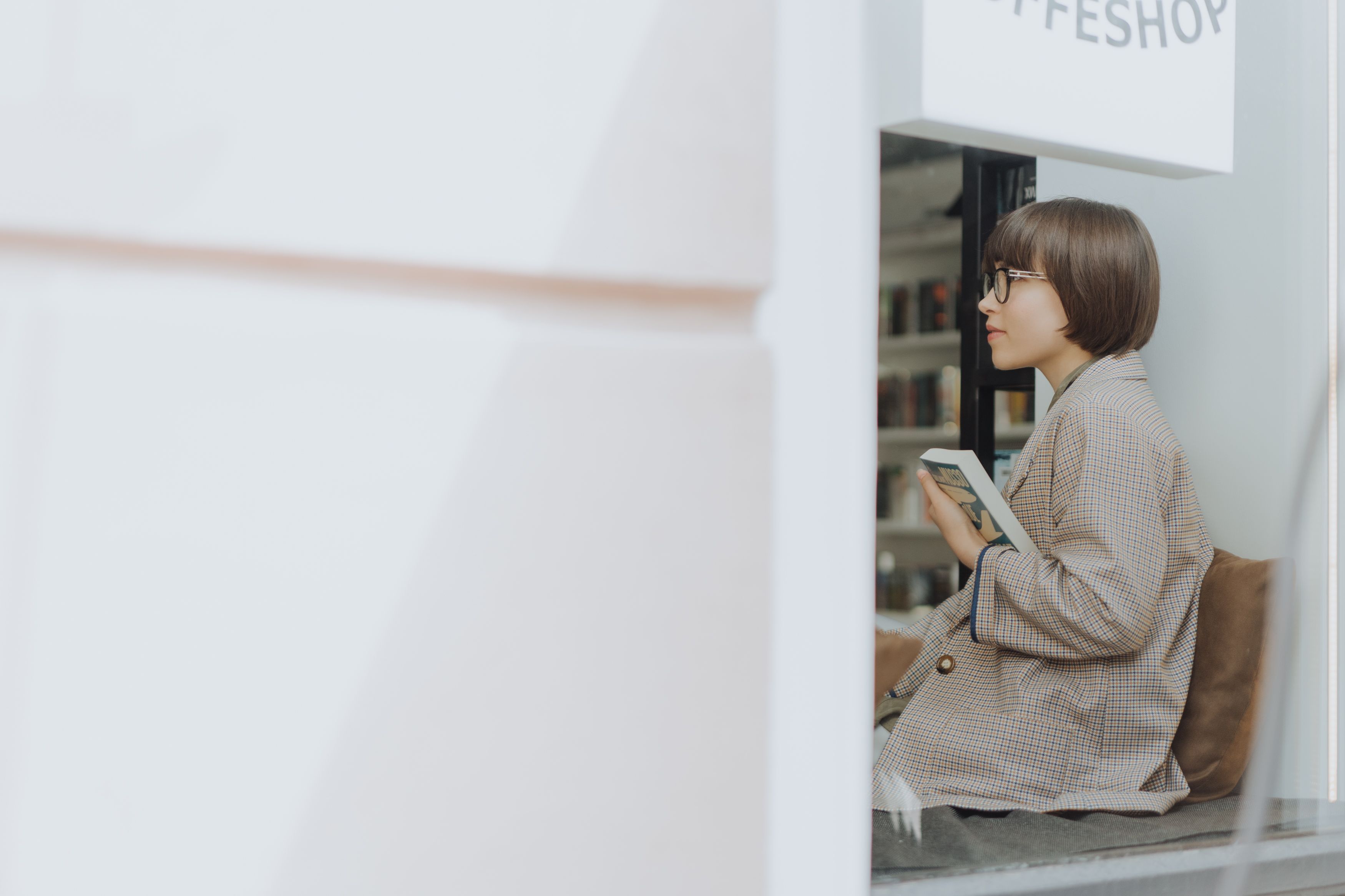 一名年轻女子穿着西装外套，坐在咖啡店的窗前看书
