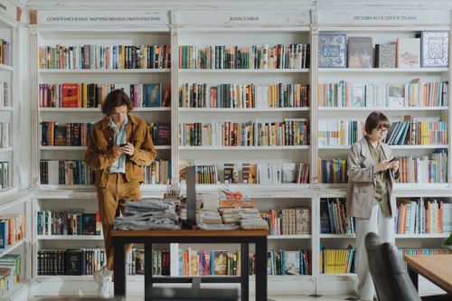 Free Two individuals browsing books in a contemporary bookstore, using cellphones. Stock Photo
