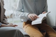 Man in White Dress Shirt and Brown Pants Sitting on White Chair Reading Book