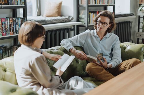 Free Two young adults enjoying a cozy moment reading and discussing books in a library. Stock Photo