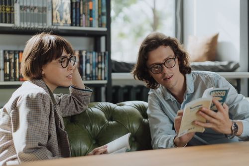 Free Two young adults in a library discussing books on a comfortable green sofa. Stock Photo