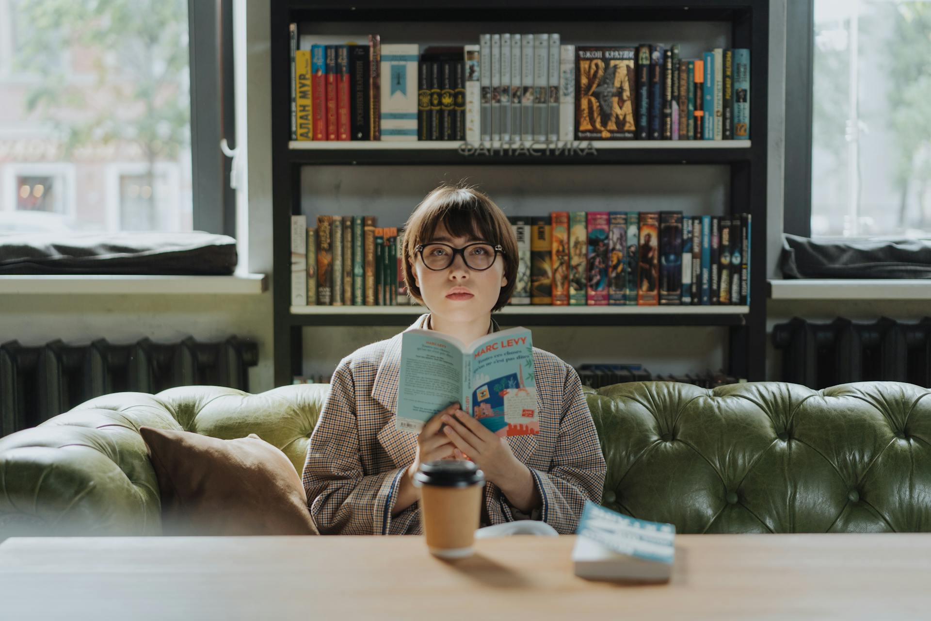 A young woman enjoys reading in a cozy library, holding a book with a coffee on the table.