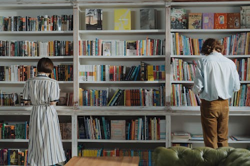 Man in White Dress Shirt Standing Beside Brown Wooden Book Shelf