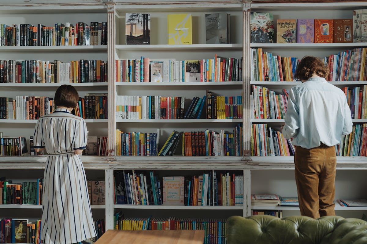 Young person sitting on the floor by a bookshelf reading a paperback book