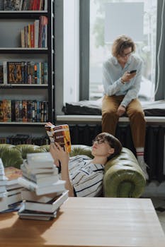 Two young adults in a cozy library setting, one reading and another browsing on a phone by the window.