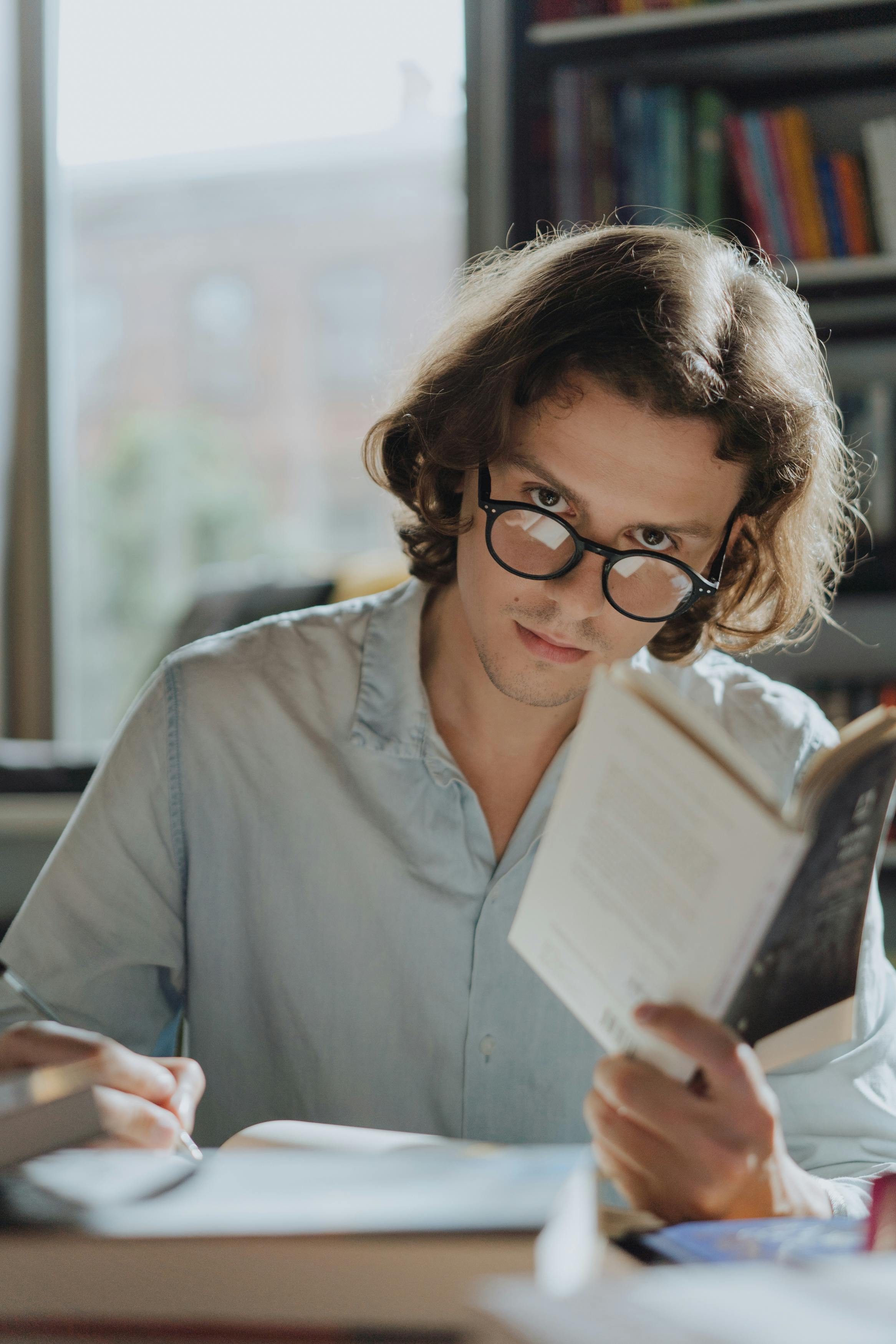 A young man with glasses reading and taking notes in a cozy library setting.