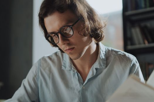 A young man wearing glasses absorbed in reading inside a serene library environment.