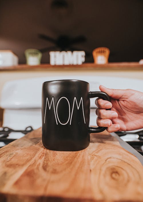 Free Crop anonymous person taking black ceramic mug with white word Mom placed on wooden table in living room Stock Photo