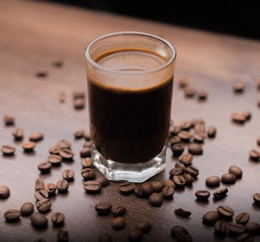 A close-up of a rich espresso in a glass surrounded by coffee beans on a wooden table.