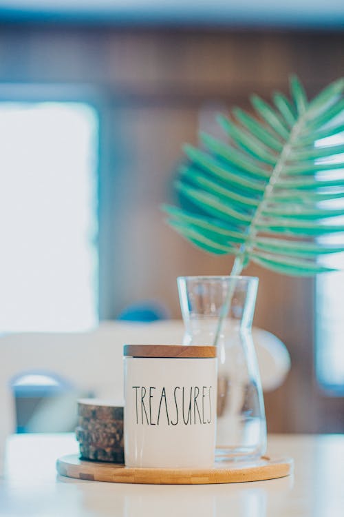 Composition of white ceramic sugar bowl with word Treasures placed on wooden board near green plant twig in glass vase on dining table